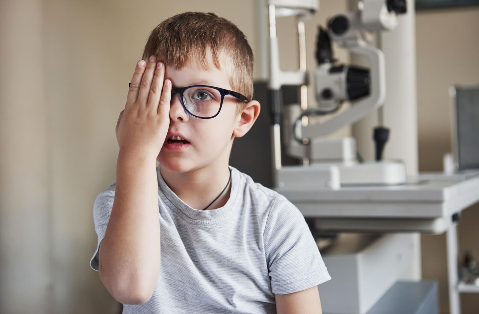 Young boy covering one eye during a vision test at the optometrist, sitting next to eye exam equipment in the clinic.