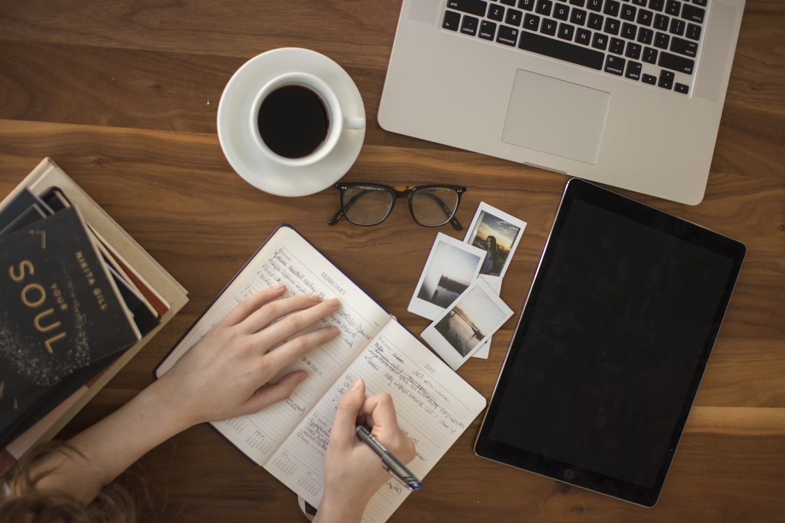 A woman is writing a problem statement in a notebook on a table, surrounded by a tablet, coffee, laptop, a few books, images, and eyeglasses