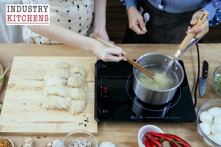 Two cooks preparing noodles on an induction cooker, with a bowl of freshly cooked noodles nearby in a commercial kitchen.