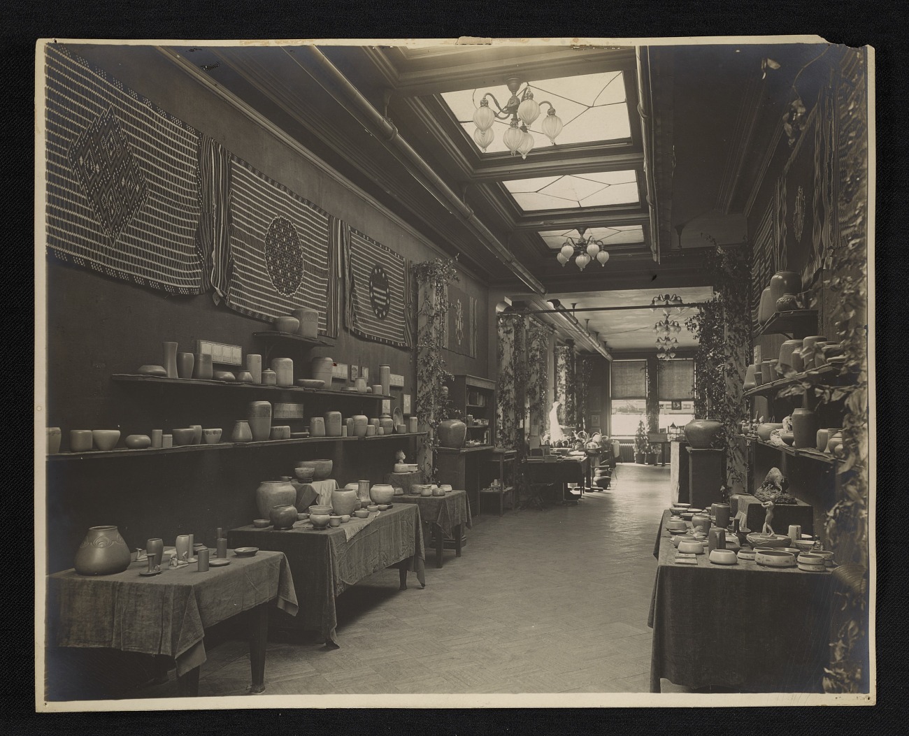 Early 20th-century pottery exhibition room featuring shelves and tables filled with handcrafted ceramic vases, bowls, and cups, with decorative textiles on the walls, natural light from skylights, and vintage lighting fixtures, showcasing Arts and Crafts movement interior design.
