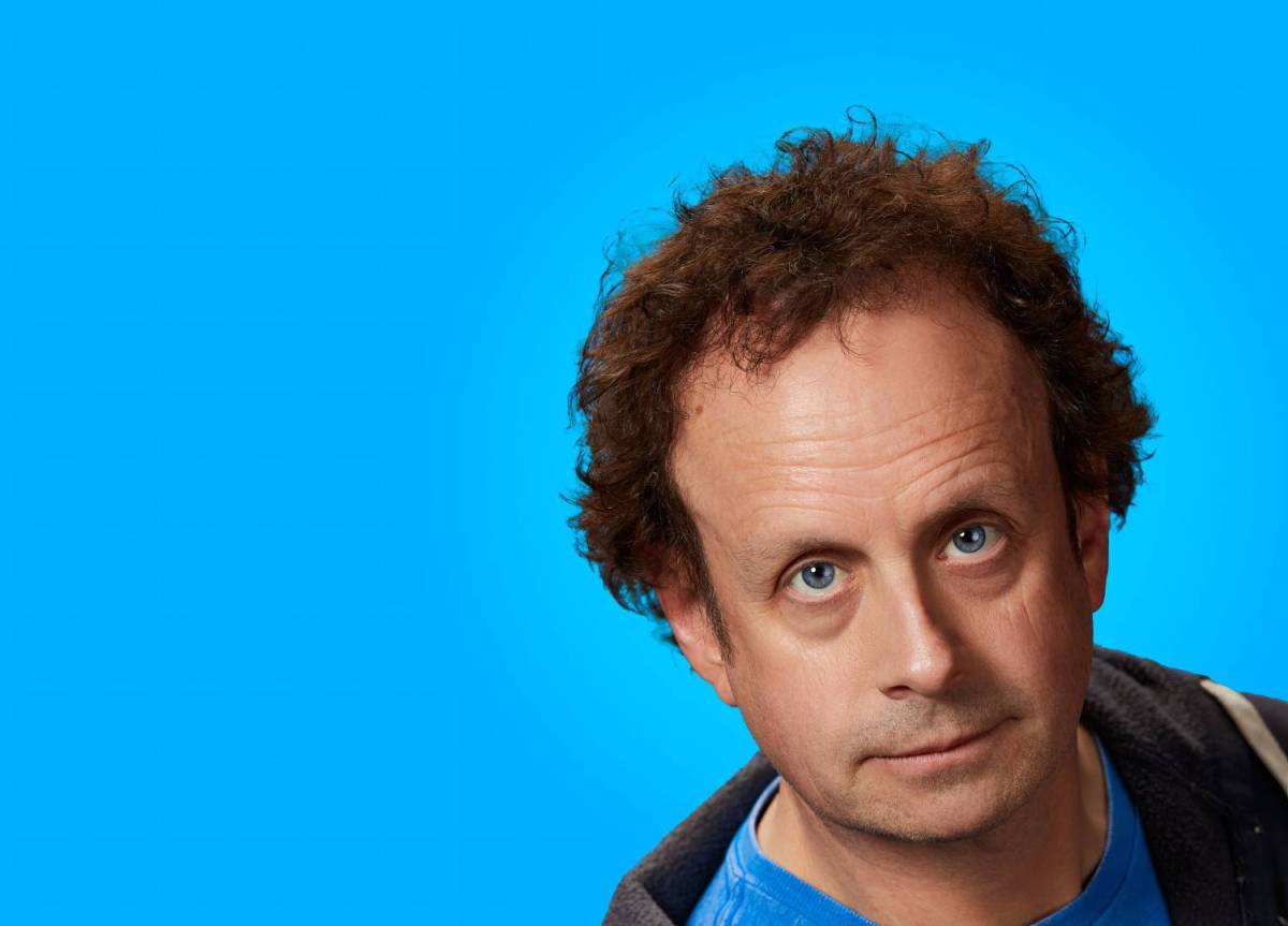 A middle-aged white man with curly hair and blue eye looks at the camera, agaisnt a bright blue background