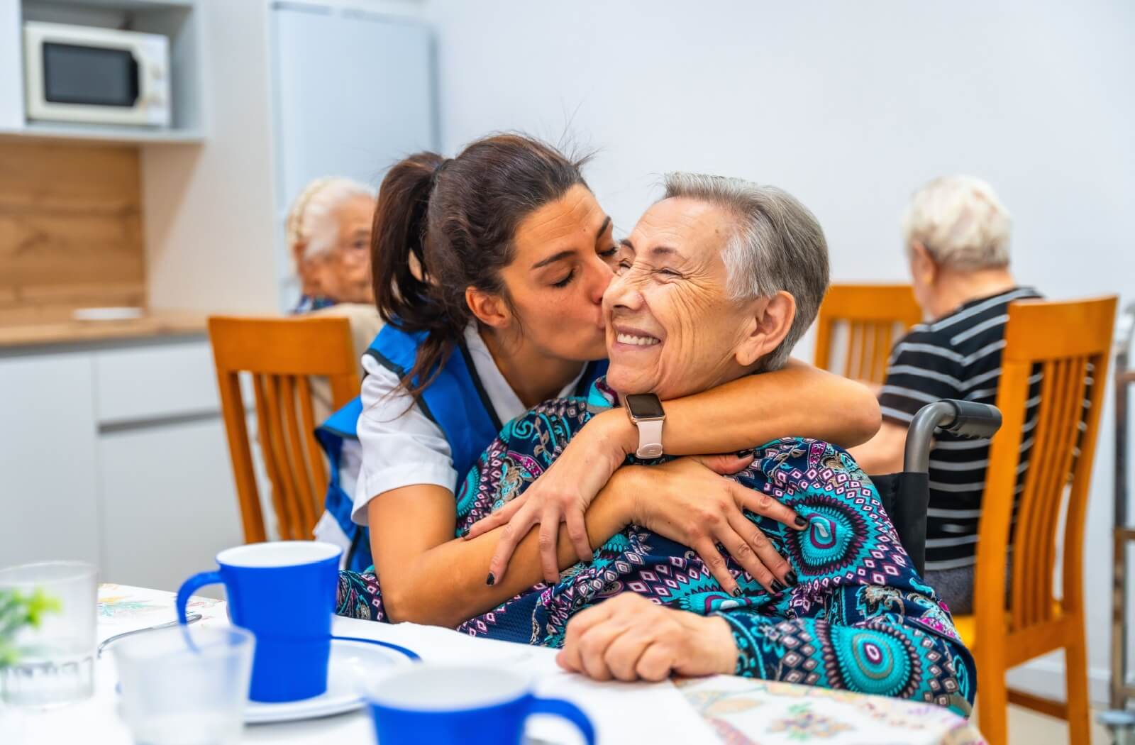 A memory care nurse hugs a senior with dementia and kisses them on the cheek during breakfast at a memory care community