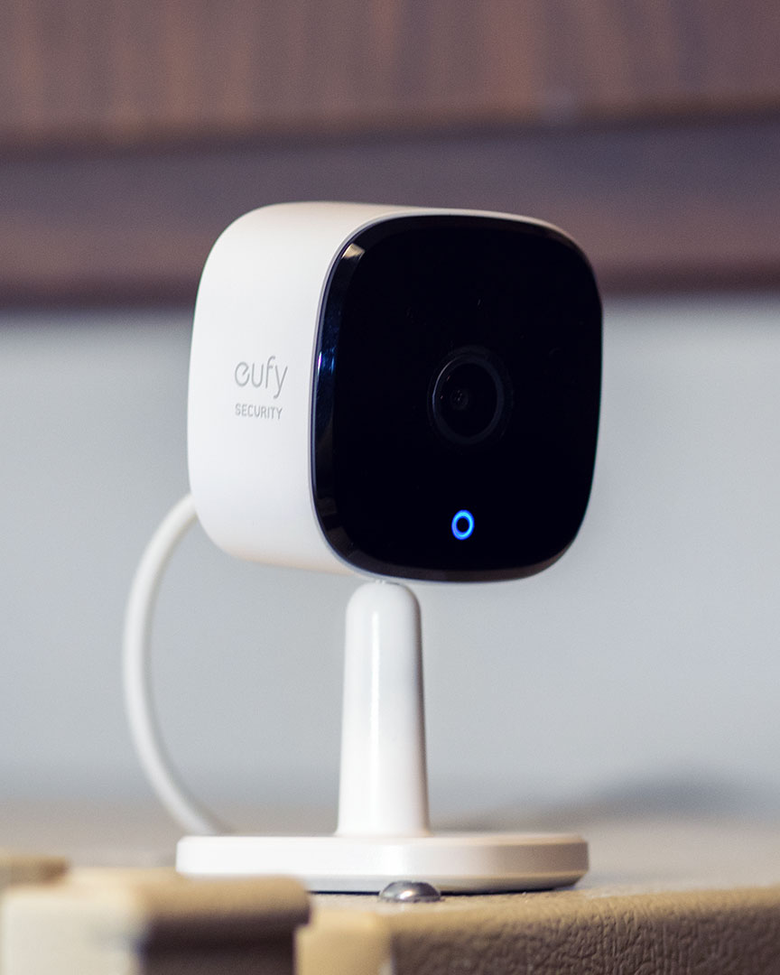 How to reset eufy Indoor Camera: A close-up of a white Eufy indoor security camera on a light surface, featuring a black lens panel and a blue indicator light. The camera is positioned on a white stand, with a cord attached.