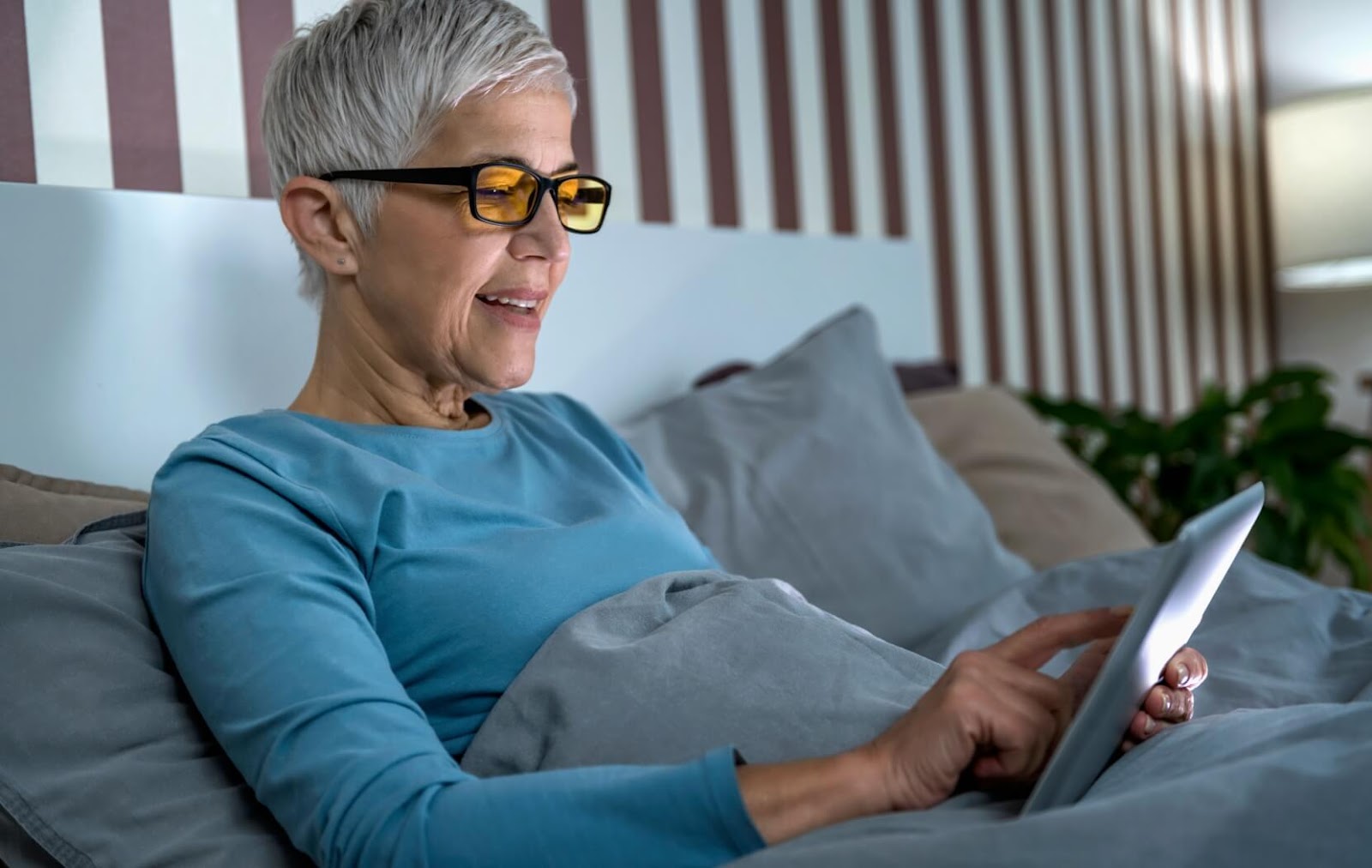 A person is wearing blue light glasses while they sit in their cozy bed and read a book on their tablet during the evening.