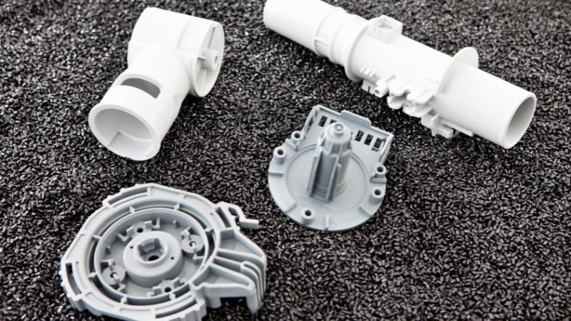 Four white and gray plastic components on a dark textured surface, likely appliance or machine parts.