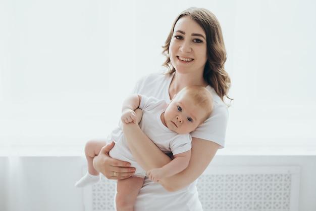 A happy young mother and a newborn baby smile hug and kiss on a white isolated window background a loving family at home