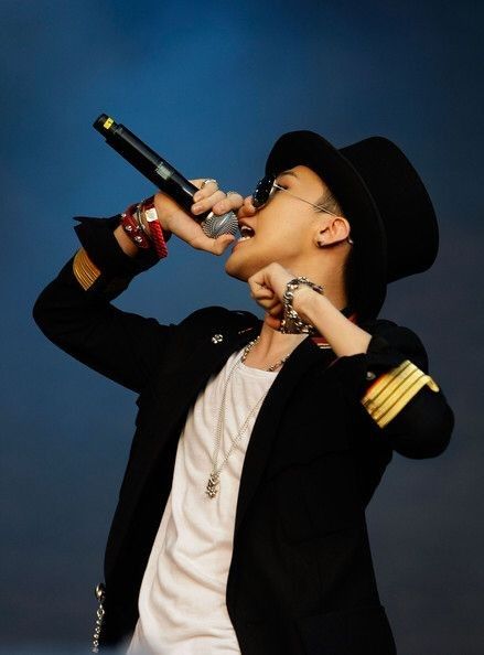 This contains an image of G-Dragon  in a black jacket and hat is holding a microphone up to his mouth