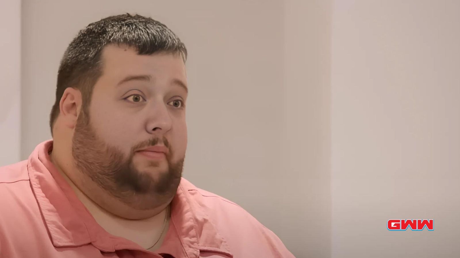 Scott in a pink shirt listens attentively in a minimalist room.