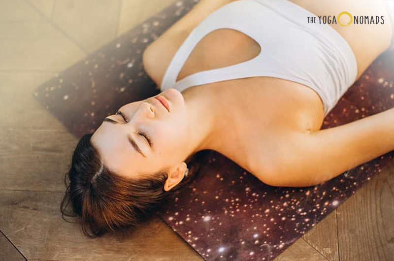 A woman laying on her yoga mat in a white yoga top, her eyes are closed indicating that she is in a corpse yoga pose.