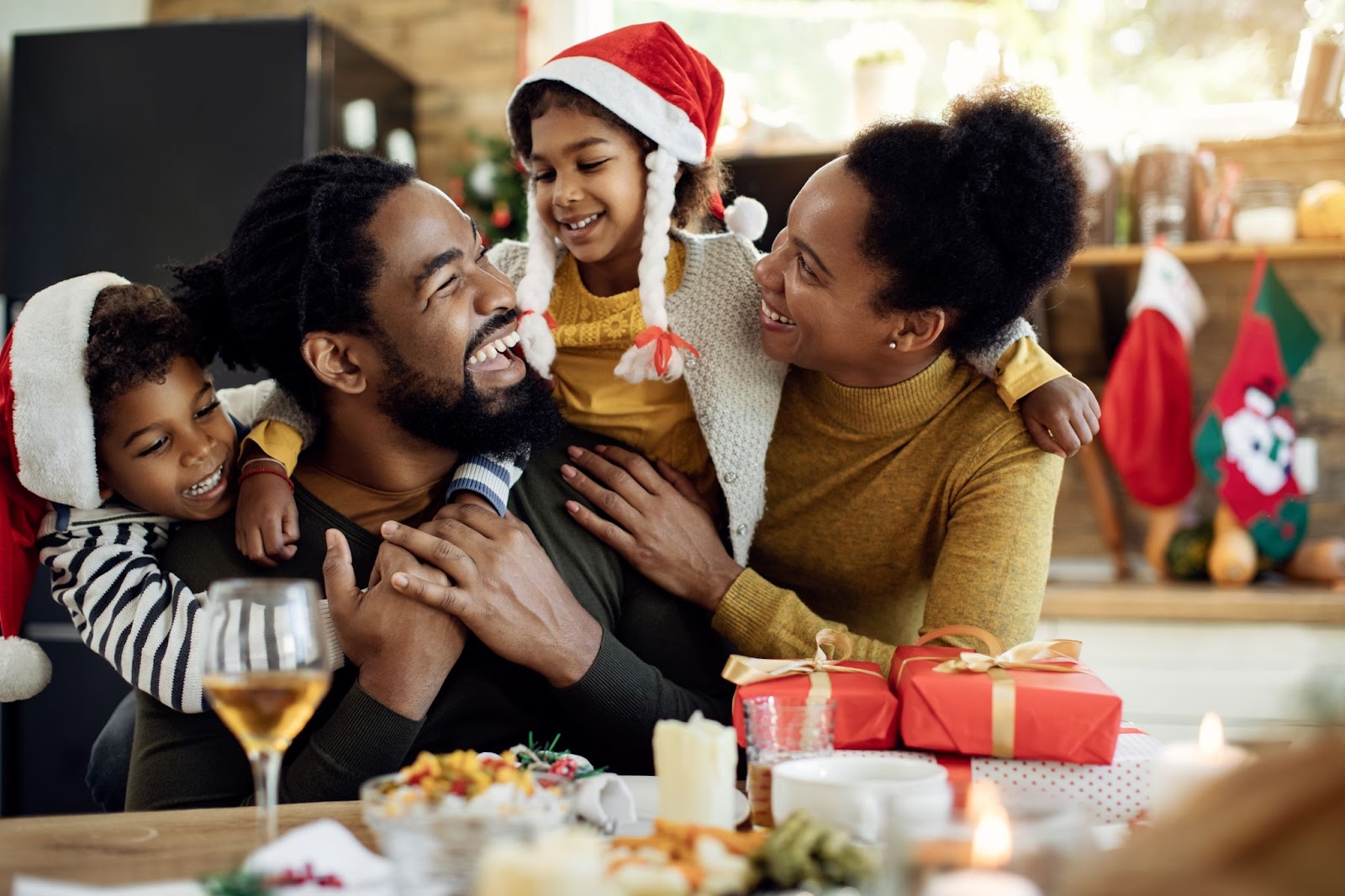 Uma família comemora feliz o Natal de frente para uma mesa posta com a ceia de Natal e presentes.