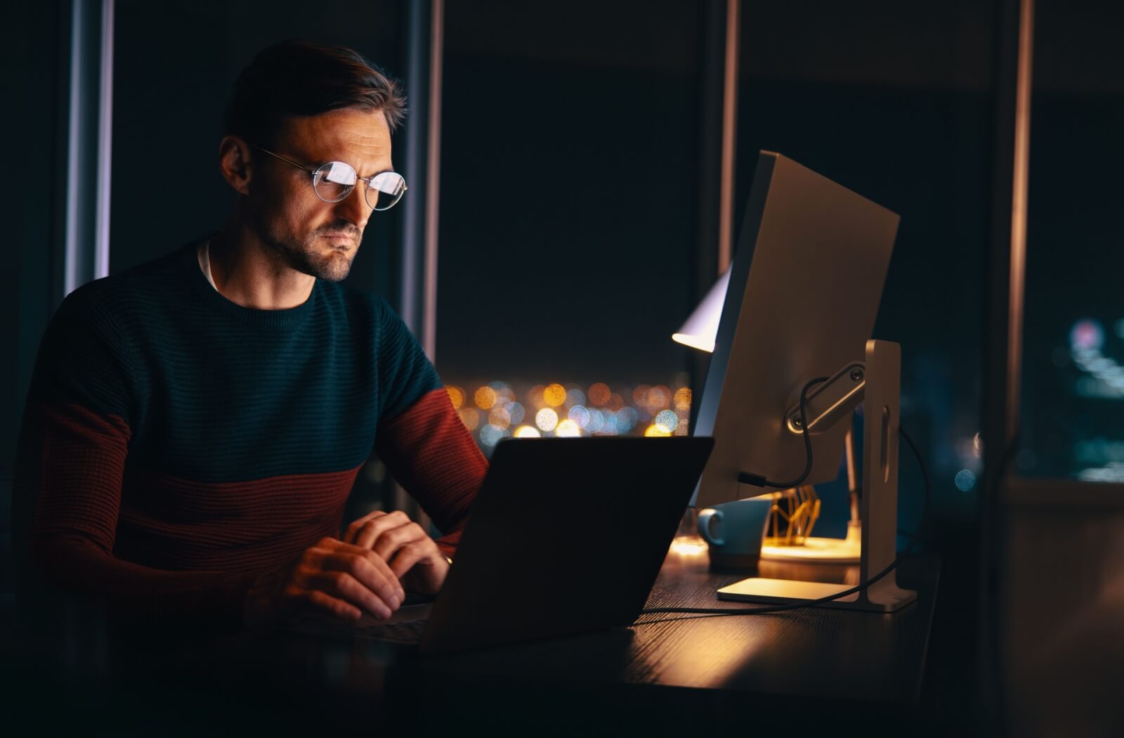 A young professional uses blue-light eyeglasses while working on multiple computers at the office late at night