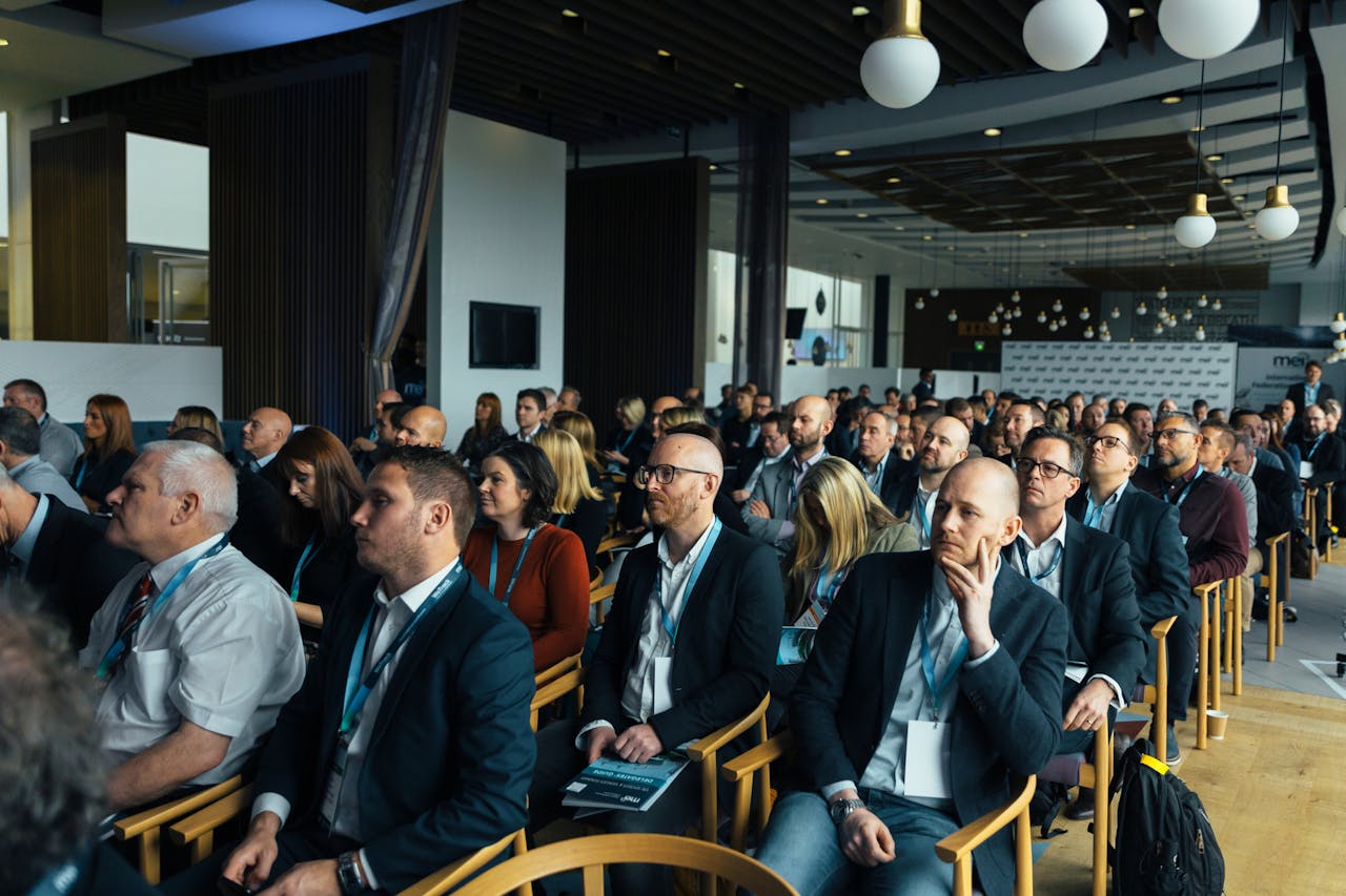 A large audience of professionals attentively listening at a conference, featuring diverse men and women dressed in business attire in a modern venue with wooden chairs and stylish lighting.