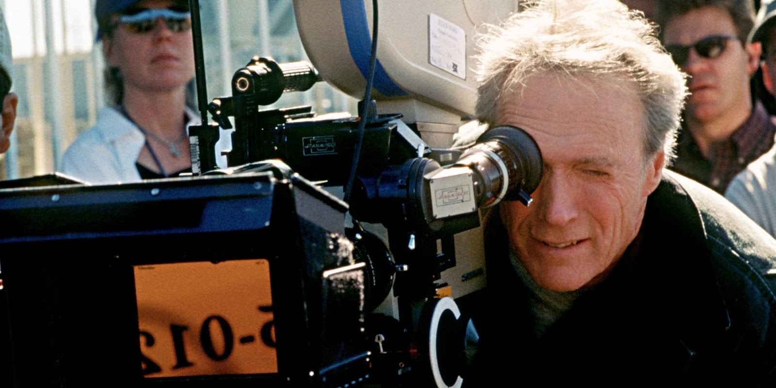 Clint Eastwood mirando por el visor de una cámara de cine mientras dirige una escena, con una expresión concentrada. El fondo incluye un asistente de producción desenfocado.