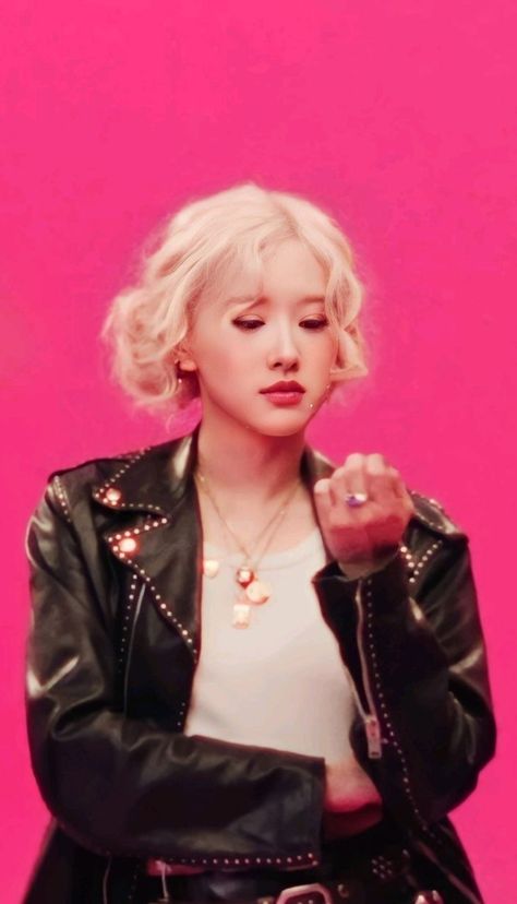 This contain Rosé with blonde hair wearing a black leather jacket and white t - shirt, standing in front of a pink background