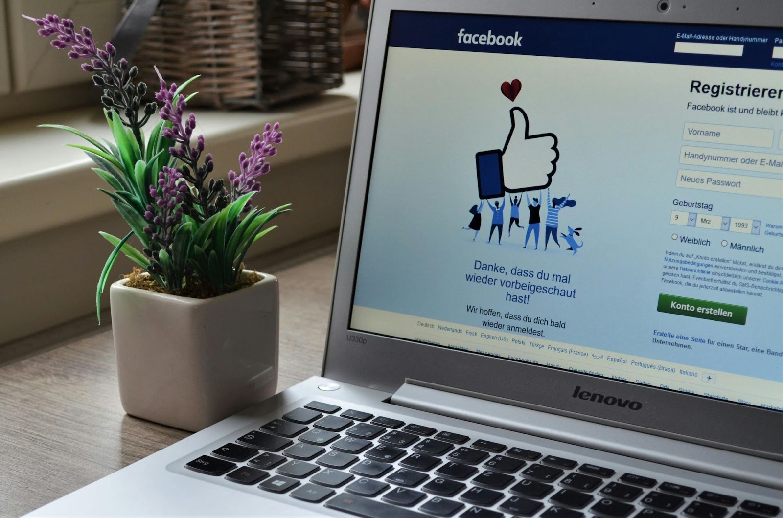 Login screen on a Lenovo laptop displaying Facebook's welcome page, with a potted plant beside it on a wooden desk