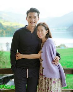 This contain Hyunbin Son Yejin  standing next to each other in front of a lake with mountains behind them