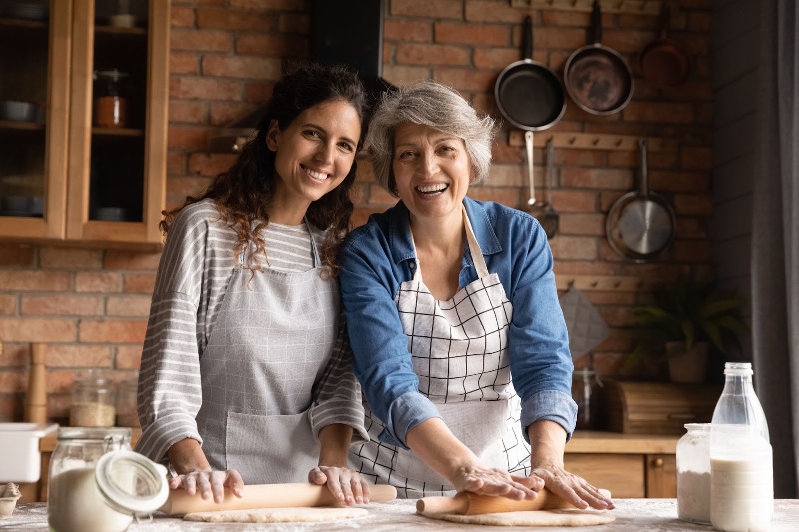  Smiling adult child and mother rolling pastry dough together in the kitchen.