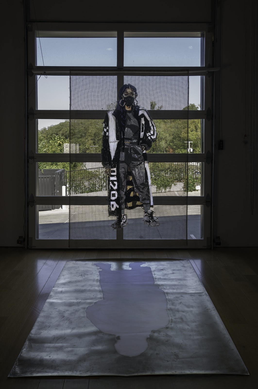 Image: Front-facing installation view of Kandy G Lopez’s Br00klynBetty and Invisible Br00klynBetty. Br00klynBetty is hung on the gallery’s glass garage door, with Invisible Br00klynBetty laid on the floor in front. Br00klynBetty is a fiber sculpture of a feminine figure in a large black and white coat–with “90210” printed on the bottom right on the coat–and a dark gray jumpsuit; and Invisible Br00klynBetty is an inverted spray-painted silhouette of the sculpture in gold on yupo paper. Allegories of Inertia, Charlotte Street, September 27-November 9, 2024. Photo by E.G. Schempf.