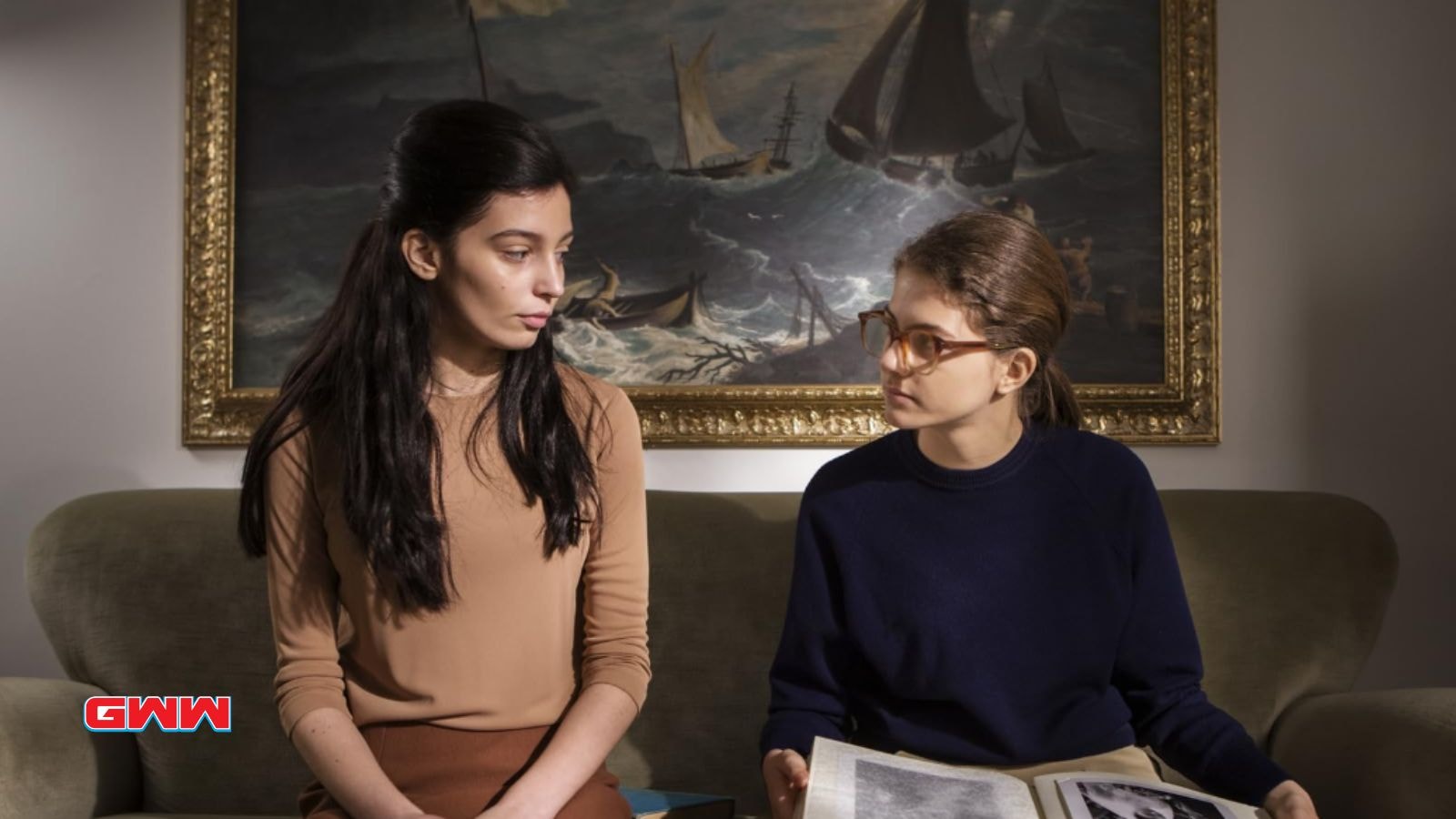 Elena and Lila converse on a couch, one holding an open book.