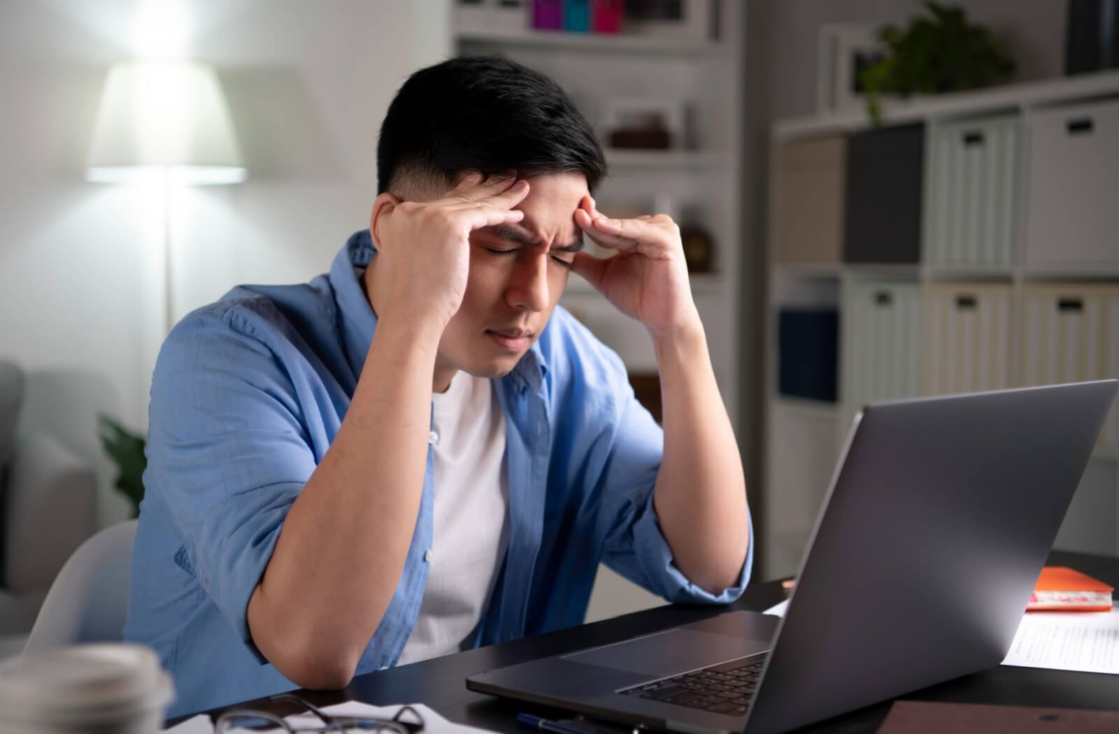 A young person with digital eye strain is sitting in front of a laptop while squinting and rubbing their head in pain.