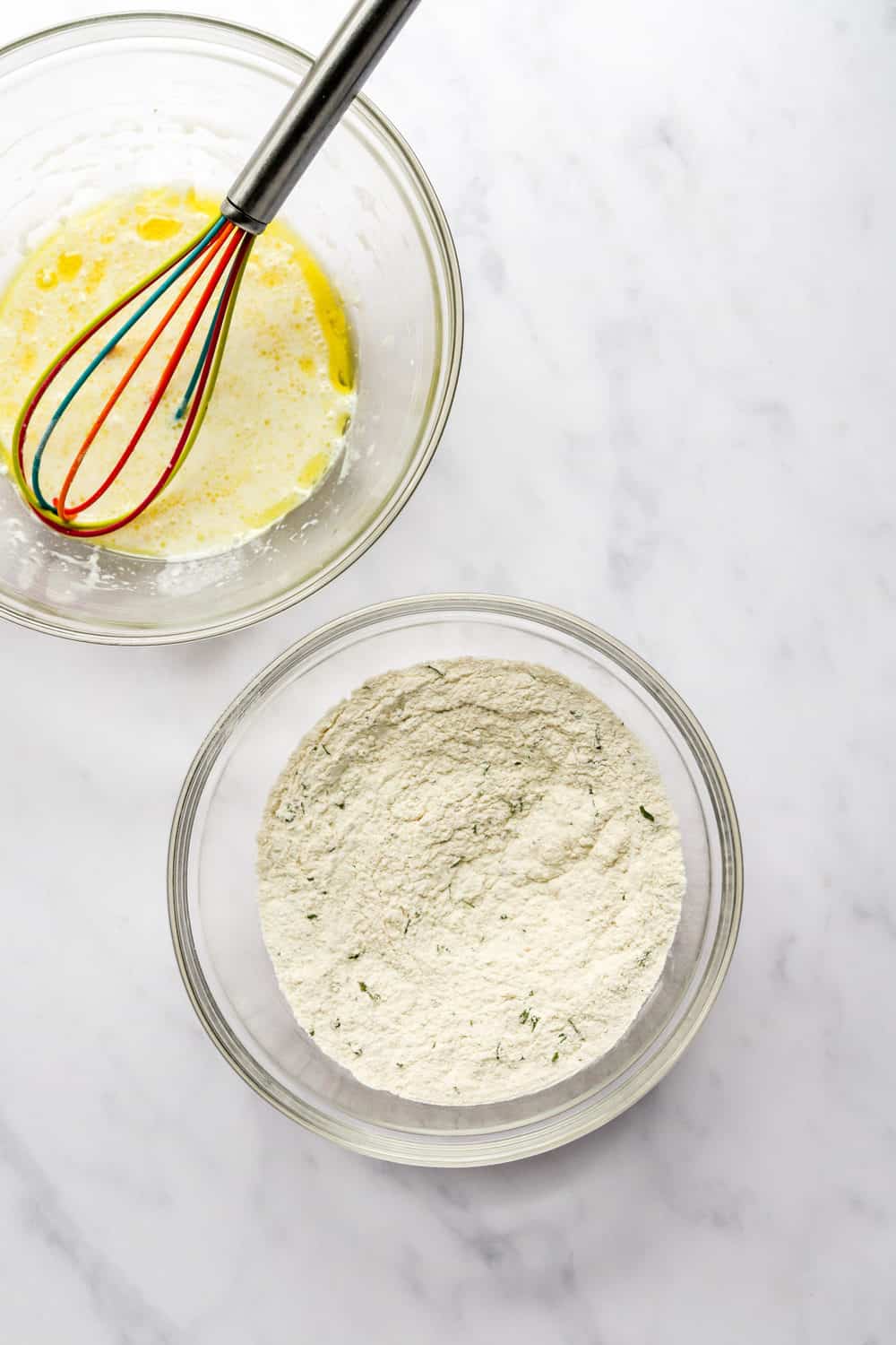 Two glass mixing bowls with one filled with dry flour ingredients and the other filled with wet ingredients with a whisk in the bowl.