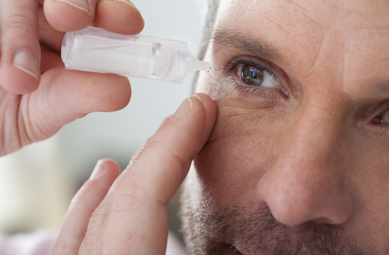 The image shows a closeup shot of a person putting eye drops in their eye to manage their dry eye symptoms.