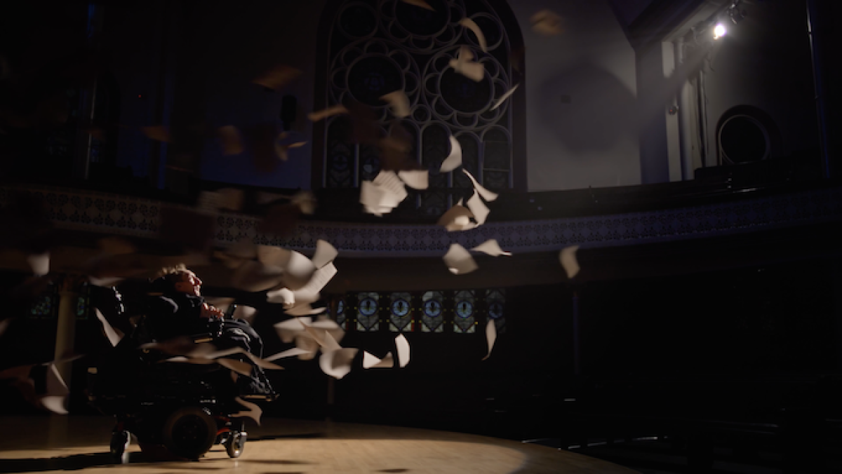 A still from dcuentary Ari Thmshwig pages flying through an empty theatre, with a composer who uses a wheelchair onstage