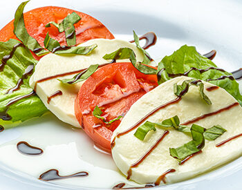 A Caprese salad featuring fresh mozzarella, sliced tomatoes, and basil, drizzled with balsamic glaze on a white plate with leafy greens.