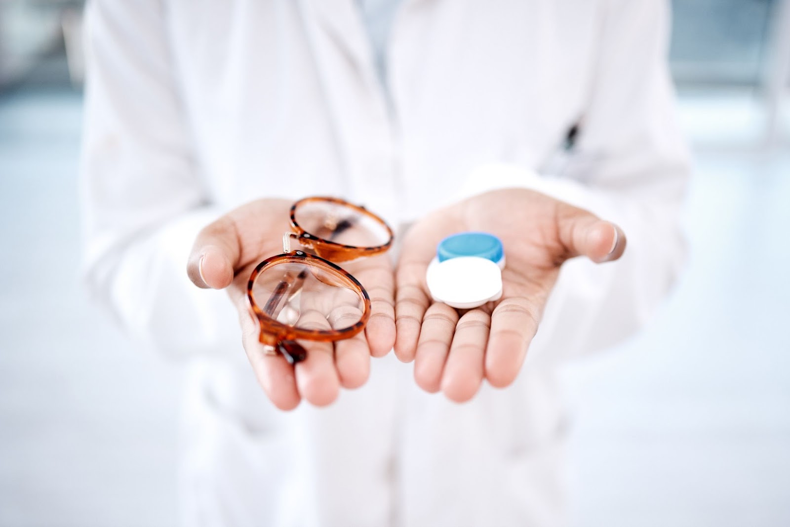 An optometrist holds their hands out to the camera, with a pair of glasses in one hand and a contact lens case in the other.