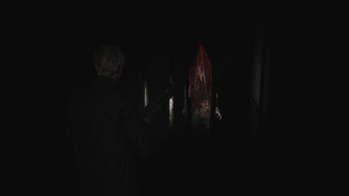 Cuplikan layar dari Silent Hill 2 Remake: James berdiri di koridor di Apartemen Blue Creek. Sesosok makhluk berdiri di depannya, di balik jeruji, menatap... meskipun kepalanya berbentuk piramida berkarat dan tidak memiliki mata.