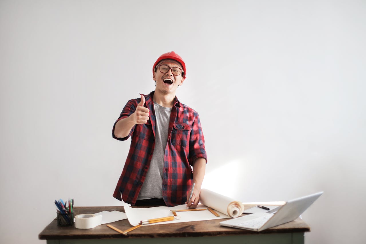 Joyful young man in a red cap and plaid shirt giving a thumbs up at his creative workspace with a laptop showing how to create online courses, papers, and drawing tools, exuding enthusiasm and positivity.