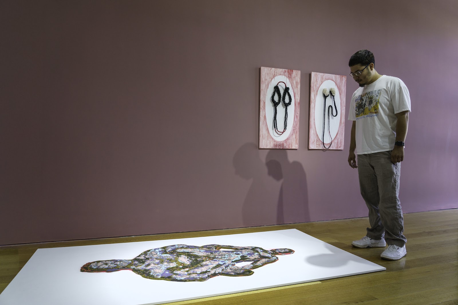 Image: Gallery view of Hugo Juarez-Avalos observing a floor installation, and wall installation beside them. Floor: Shawn Bitters’s Arch, dimensions variable. Hand-dyed sand and screen-printed sand sculpture of two figures engaged in intercourse. Wall: Yulie Urano’s Hair Portrait #1-7, 30” x 20” x 2” per portrait. Two pink and white painted frames with sculptures of braided hair and rope affixed in the middle of each. Allegories of Inertia, Charlotte Street, September 27-November 9, 2024. Photo by E.G. Schempf.