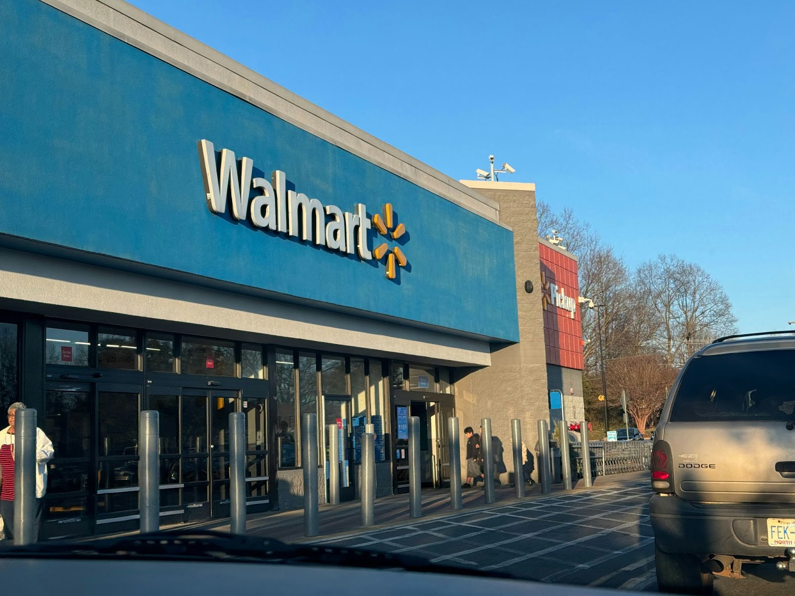 Exterior view of a Walmart store with a pickup section and customers entering, seen from a car in the parking lot.