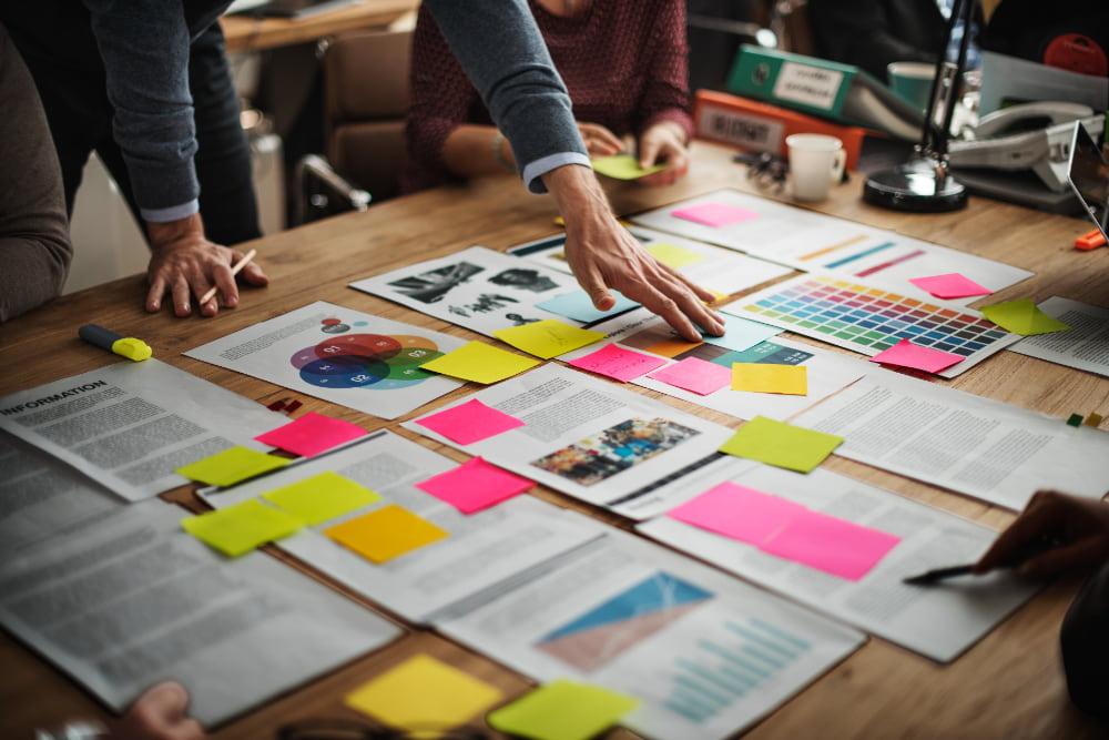 Mesa de trabalho repleta de documentos, gráficos e post-its coloridos, com várias pessoas colaborando.