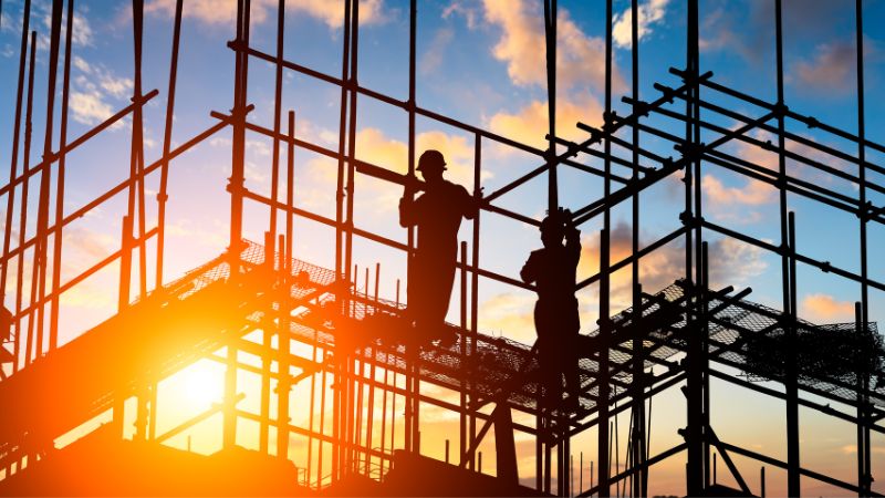 Two construction workers stand on scaffolding, silhouetted against a sunset sky.