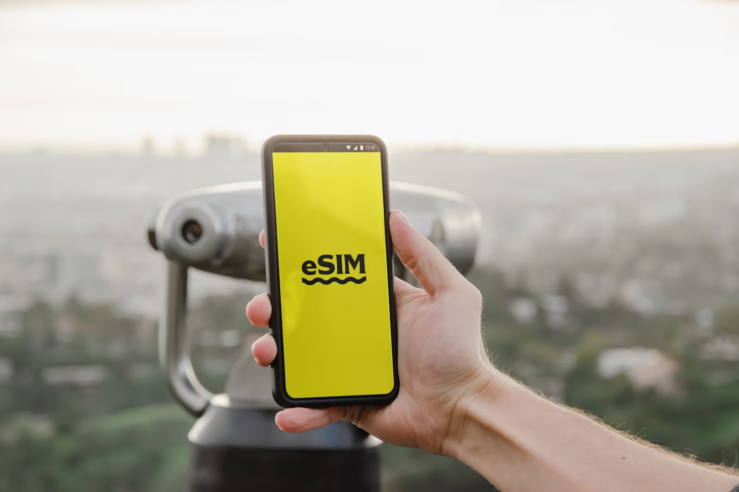 黄色い画面に「eSIM」と表示されたスマートフォンを持つ手。背景には望遠鏡とぼやけた都市の風景が広がっている。