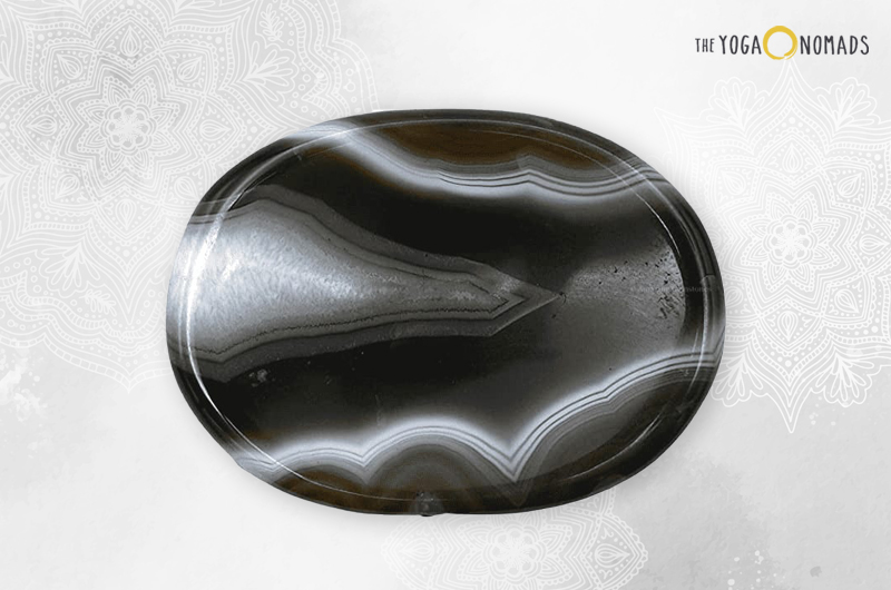 A polished black onyx stone with white and grey banding patterns. The stone is oval-shaped and has a glossy surface, reflecting light on its top left side. It is placed against a light background with subtle floral patterns around the edges.