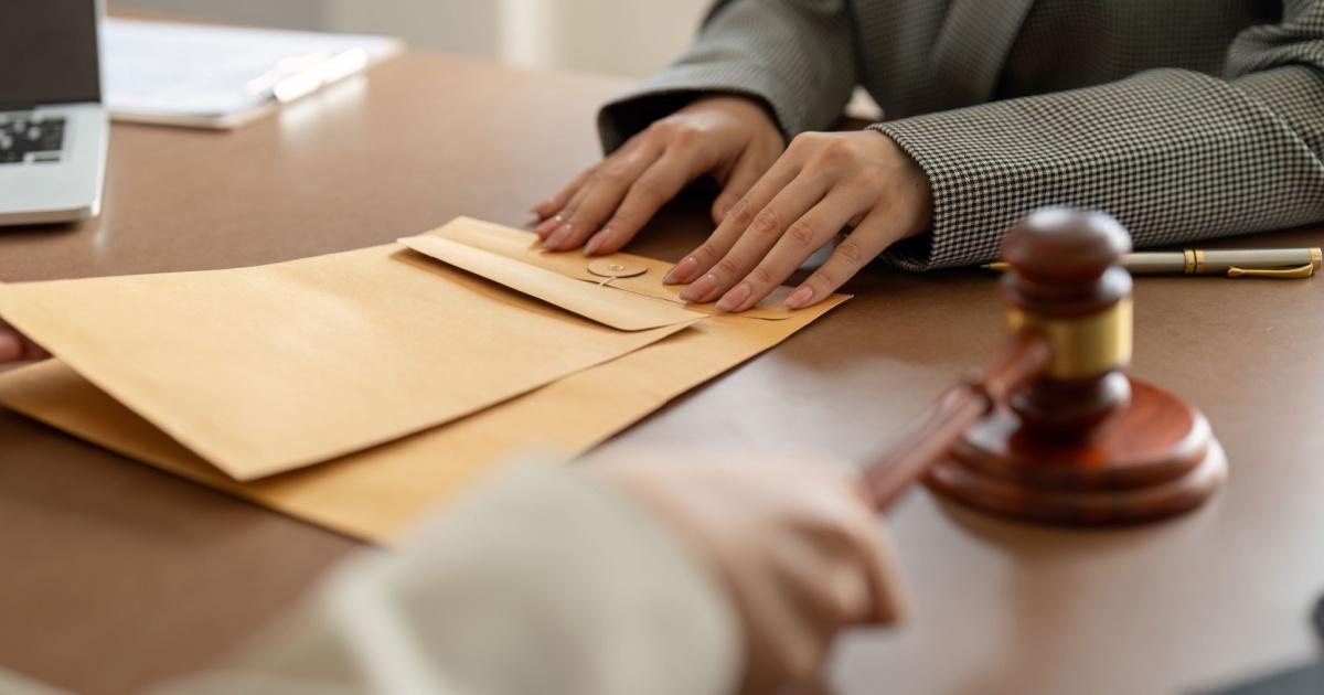 Close de uma reunião em um ambiente formal, com foco nas mãos de uma pessoa vestindo um blazer repousando sobre um envelope pardo em uma mesa. Um martelo de juiz aparece em destaque no lado direito da imagem, enquanto no fundo há um laptop e outros itens de escritório. A cena remete a um contexto jurídico ou de arbitragem.