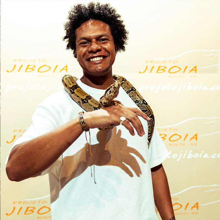 Homem sorridente segurando uma cobra, símbolo de conexão com a natureza e espiritualidade, em evento do Projeto Jiboa, destacando sua felicidade e bem-estar.