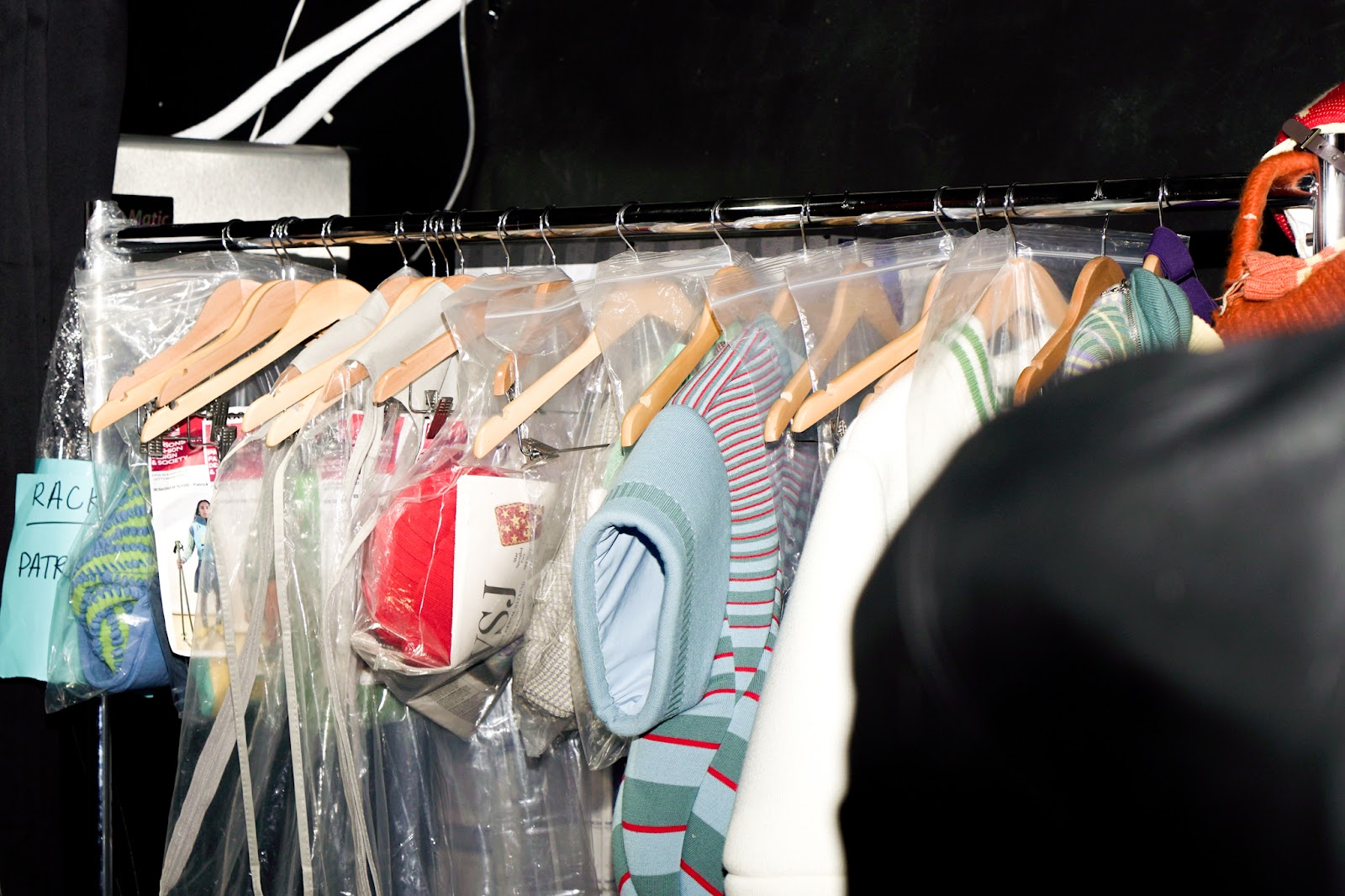 A backstage clothing rack holds garments wrapped in plastic with hangers, organized for a fashion show.