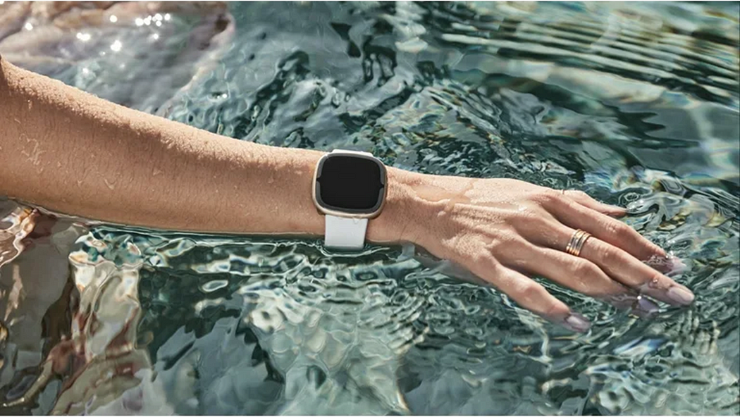 Woman wearing Fitbit Sense 2 at the swimming pool with the fitness tracker halfway submerged in water. Fitbit Sense 2 is designed to be water-resistant up to 50 meters.