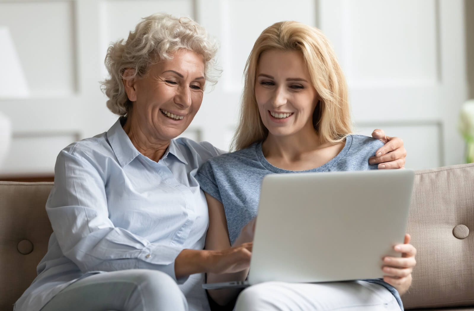 An adult child and their parent sitting on the couch in front of an open laptop researching more about dementia and smiling.