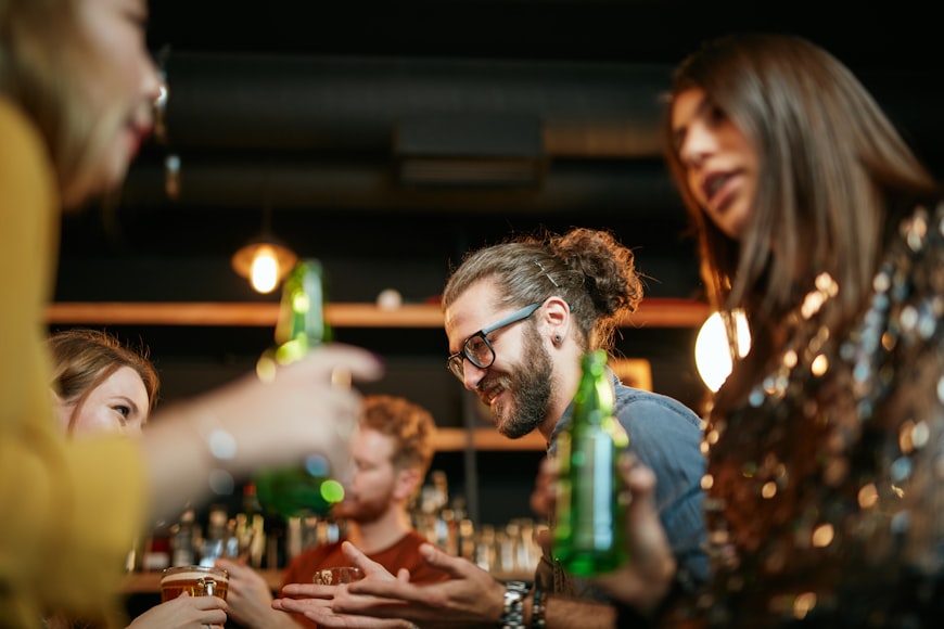 A small group of best friends standing at a pub, drinking beer, chatting and having fun. Focus on the bartender wiping the drinking glass. Nightlife.