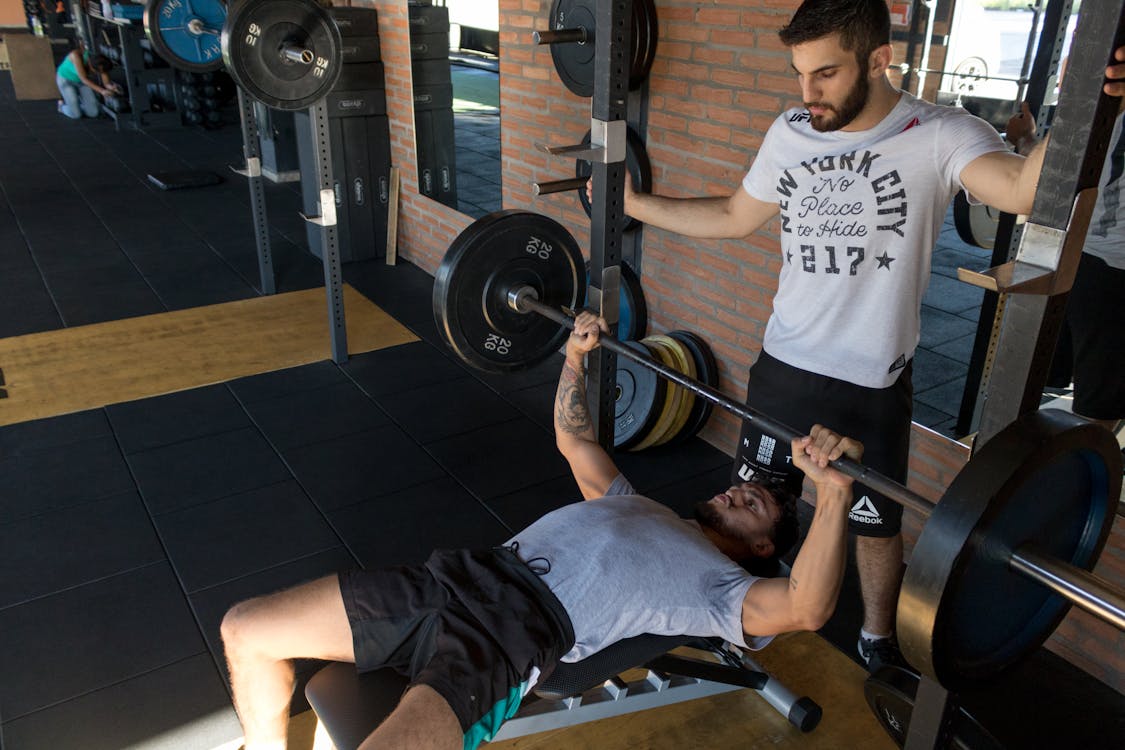 Free A strong man lifts weights on a bench while a trainer assists in a modern gym setup. Stock Photo