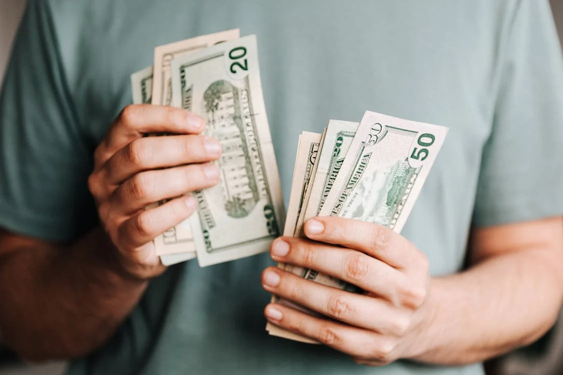 A person in a green shirt holding and counting multiple denominations of U.S. dollar bills, including $20 and $50 notes, emphasizing cash handling