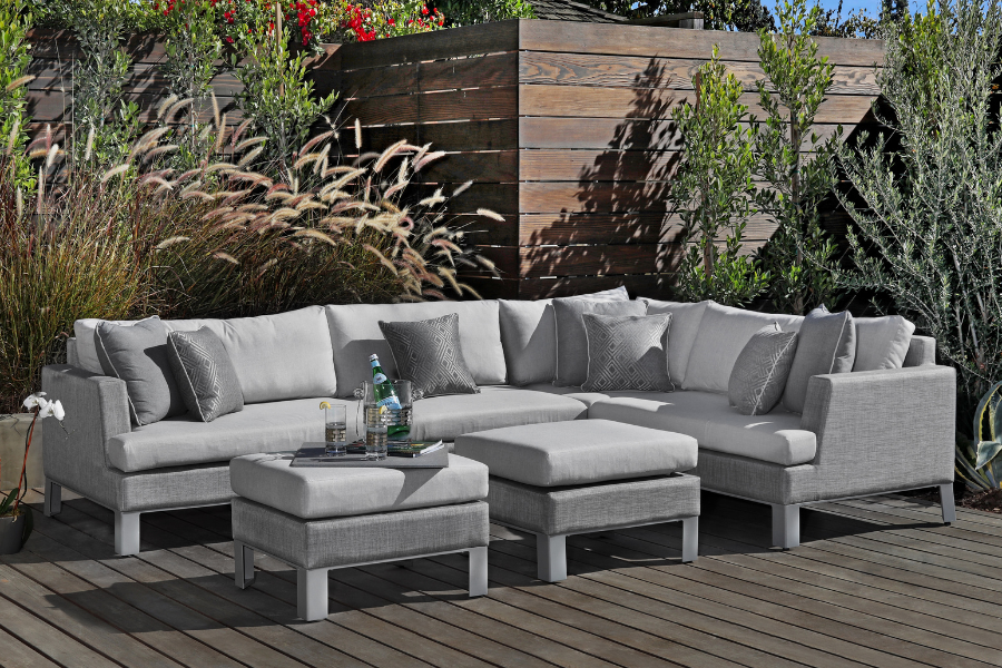 A modern outdoor sectional with gray Sunbrella® fabric, paired with matching ottomans and cushions, set on a wooden deck surrounded by lush greenery and a wooden privacy wall.