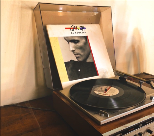 A vintage turntable playing a vinyl record with an album cover displayed behind it featuring a black-and-white photo of a man.