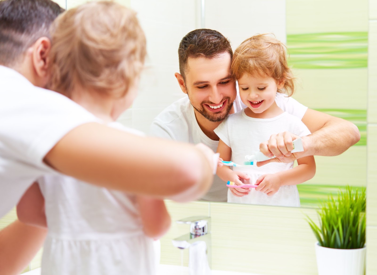 A parent demonstrates proper tooth brushing technique to his young child, both smiling as they practice good oral hygiene together.