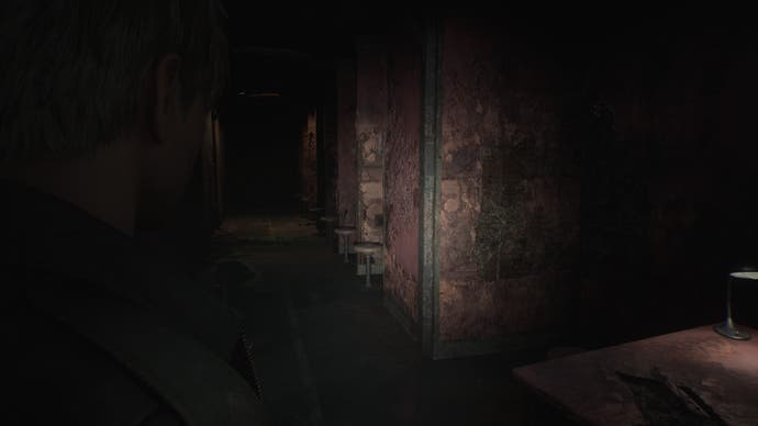 Tangkapan layar dari Silent Hill 2 Remake: James berdiri dalam kegelapan, melihat ke bawah deretan kursi di ruang kunjungan penjara yang tampak terbengkalai.