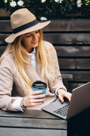 Woman in hat drinking coffee and working on laptop outside